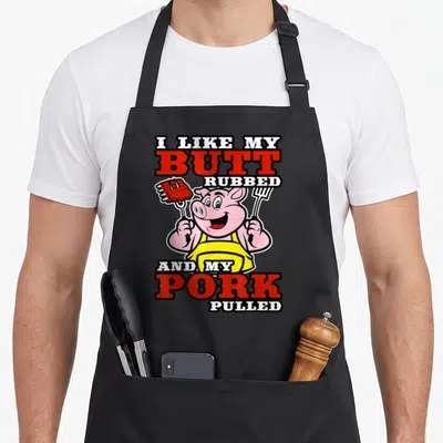 Butt barbecue apron - Cartoon pig in yellow apron holding ribs and fork with red and white stacked text — shown on a black cotton canvas apron against a white background