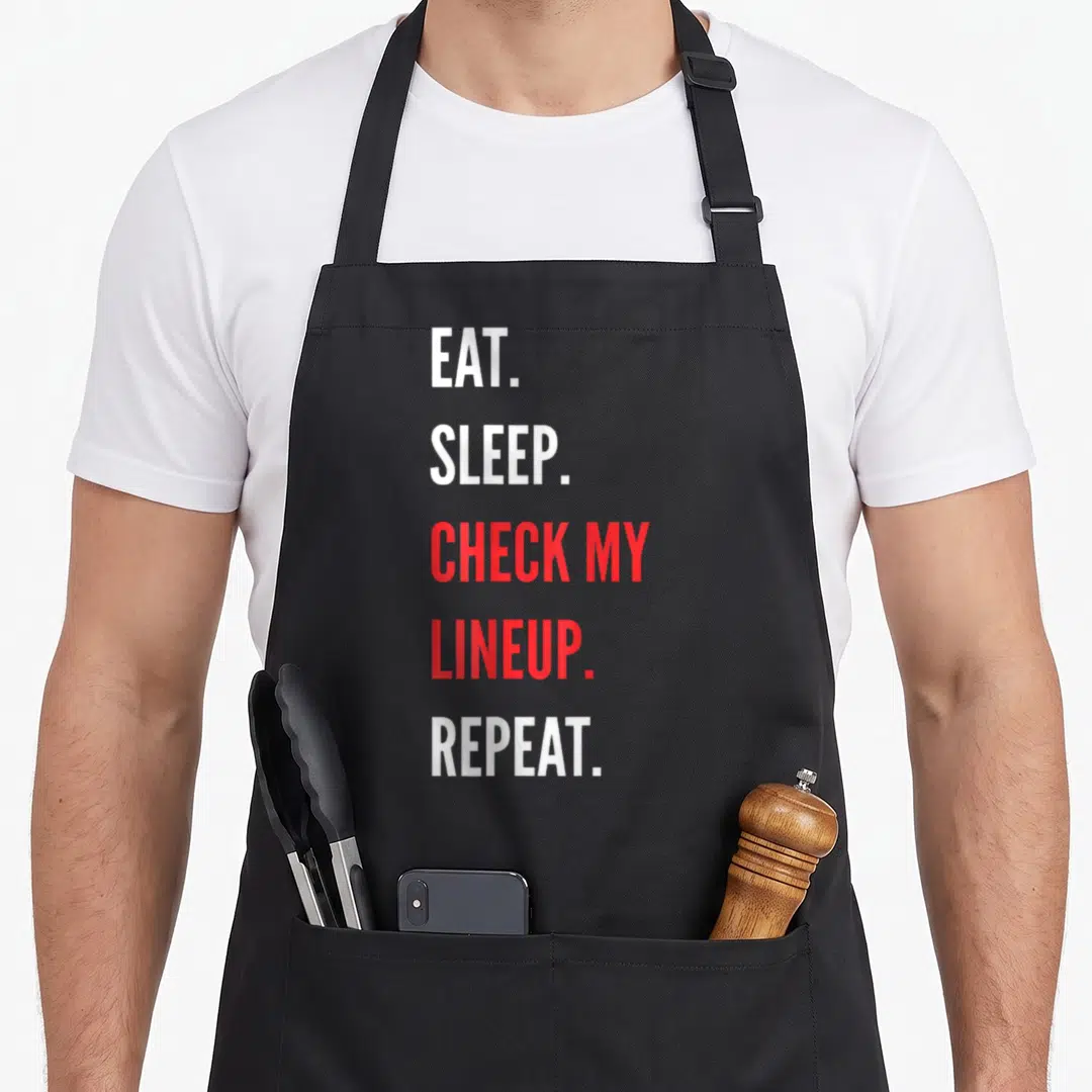 Check football apron - Vertical stack of white and red text reading EAT SLEEP CHECK MY LINEUP REPEAT — shown on a black cotton canvas apron against a white background