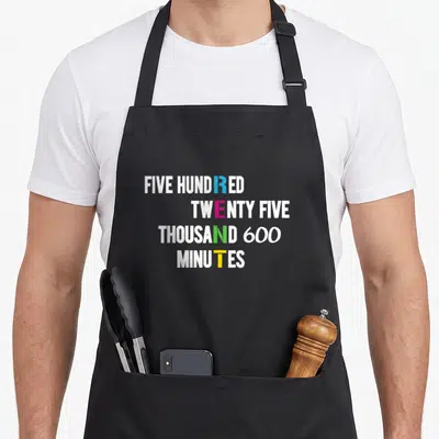 Cute minute apron - Stacked text reading 525600 minutes with blue pink green and yellow letters — shown on a black cotton canvas apron against a white background