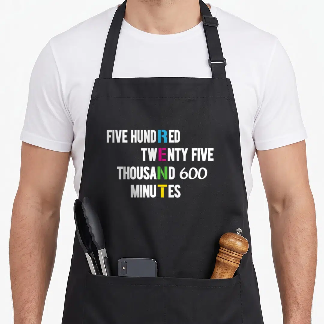 Cute minute apron - Stacked text reading 525600 minutes with blue pink green and yellow letters — shown on a black cotton canvas apron against a white background