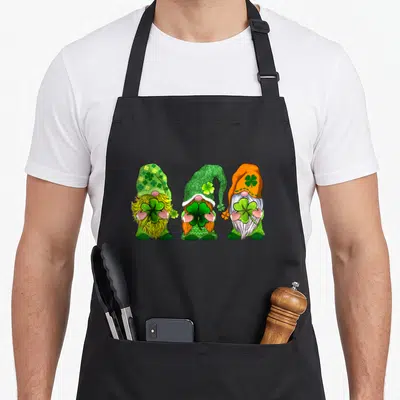 Three gnome st patrick day aprons - Three cartoon gnomes holding shamrocks in green orange yellow and white — shown on a black cotton canvas apron against a white background