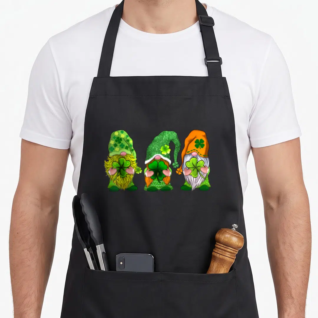 Three gnome st patrick day aprons - Three cartoon gnomes holding shamrocks in green orange yellow and white — shown on a black cotton canvas apron against a white background