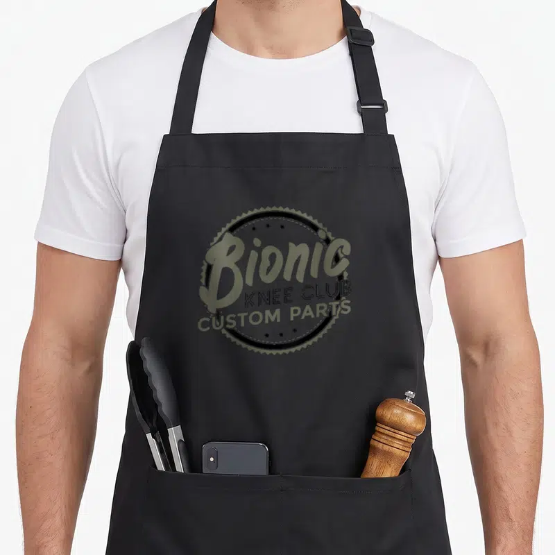 Knee replacement parts of an apron - Bionic Knee Club Custom Parts text inside a black jagged circle with olive — shown on a black cotton canvas apron against a white background