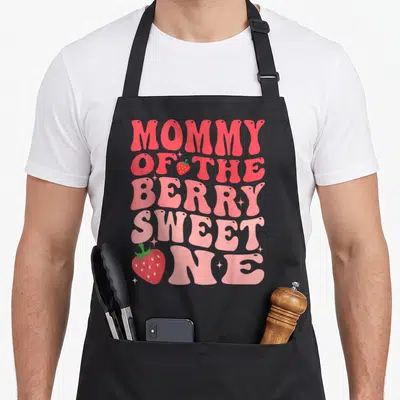 Berry sweet strawberry apron - Retro pink text Mommy of the Berry Sweet One with strawberries and stars — shown on a black cotton canvas apron against a white background
