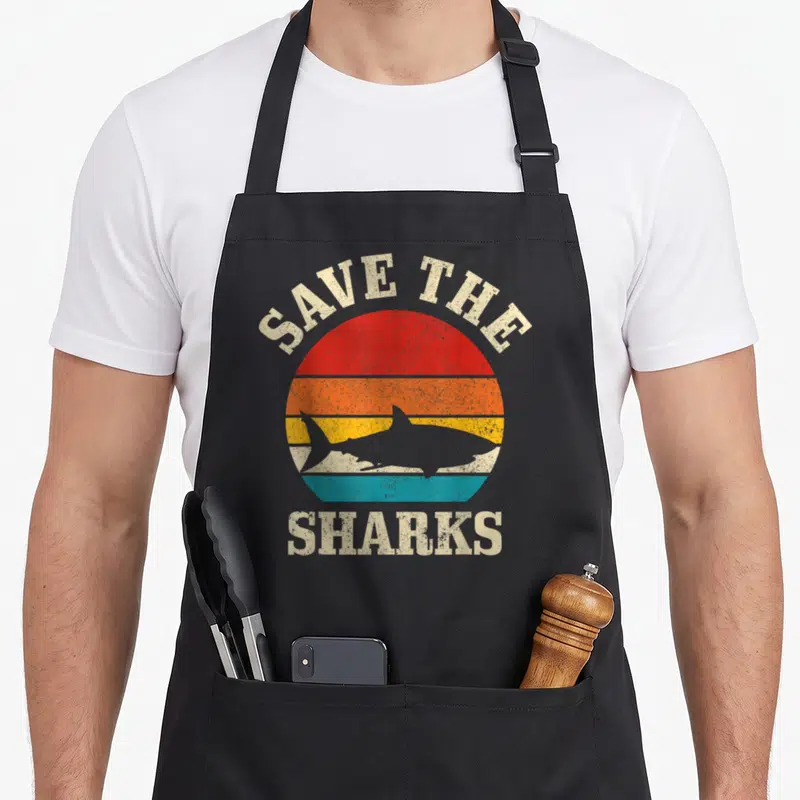 Save shark lady apron - Retro striped sun with shark silhouette and text SAVE THE SHARKS in red orange yellow teal beige — shown on a black cotton canvas apron against a white background