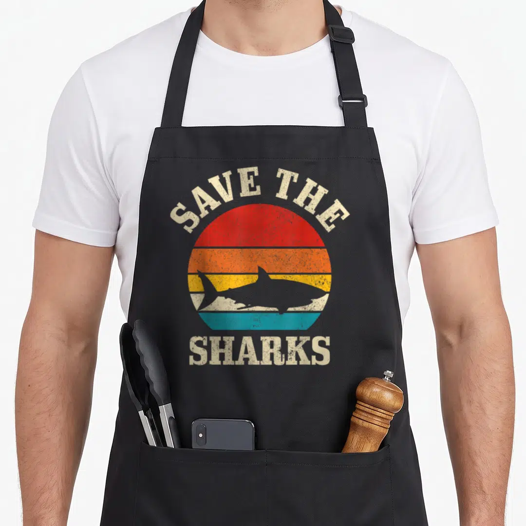 Save shark lady apron - Retro striped sun with shark silhouette and text SAVE THE SHARKS in red orange yellow teal beige — shown on a black cotton canvas apron against a white background