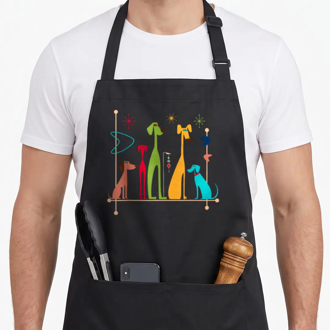 50s 60s modern apron - Five stylized dogs in orange, green, red, and blue with atomic stars on a beige line — shown on a black cotton canvas apron against a white background