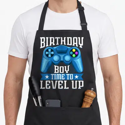 Birthday level gamer video game apron - Blue game controller with BIRTHDAY BOY TIME TO LEVEL UP text and stars — shown on a black cotton canvas apron against a white background