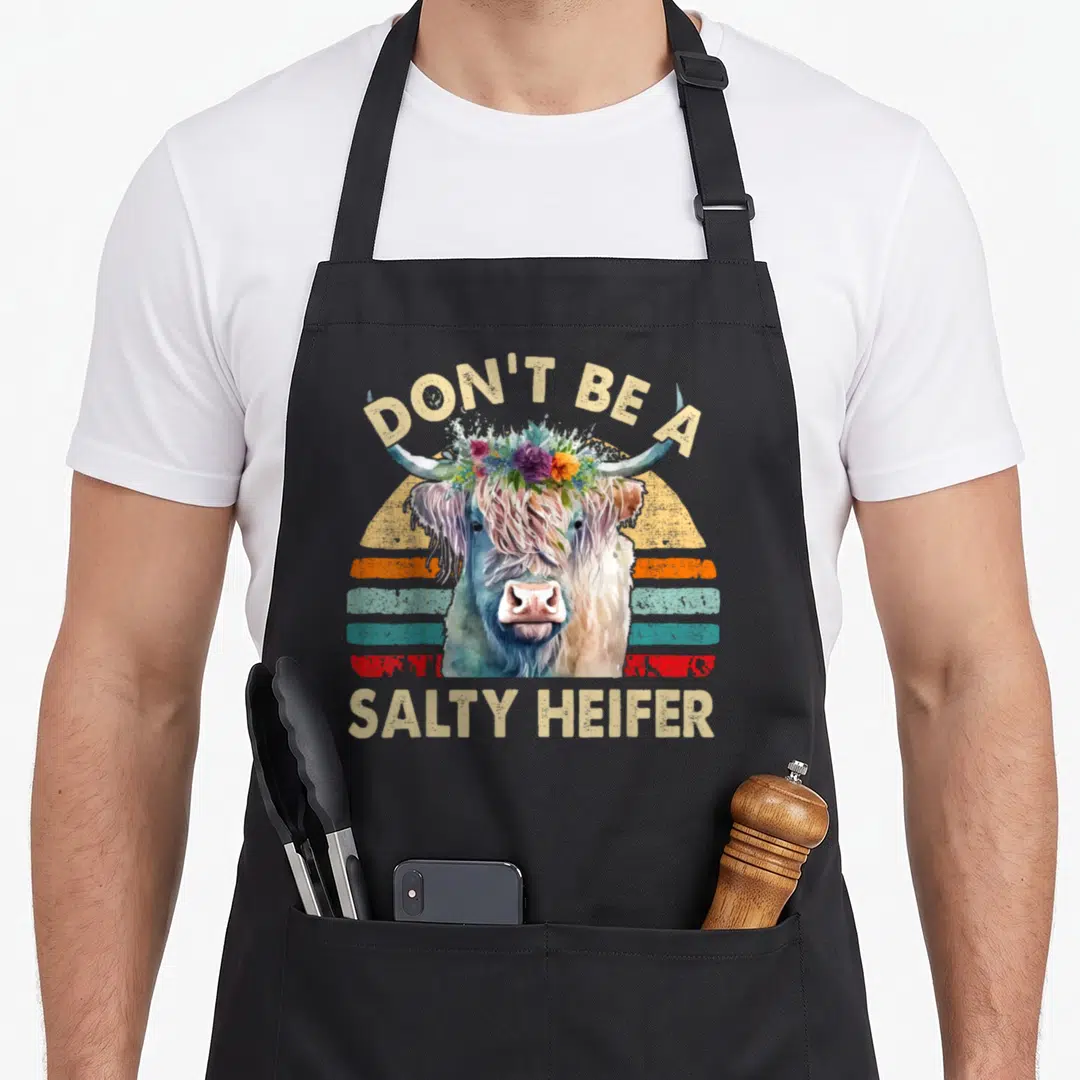 Highland cow print apron - Watercolor cow with flower crown and retro sunset stripes under salty heifer text — shown on a black cotton canvas apron against a white background
