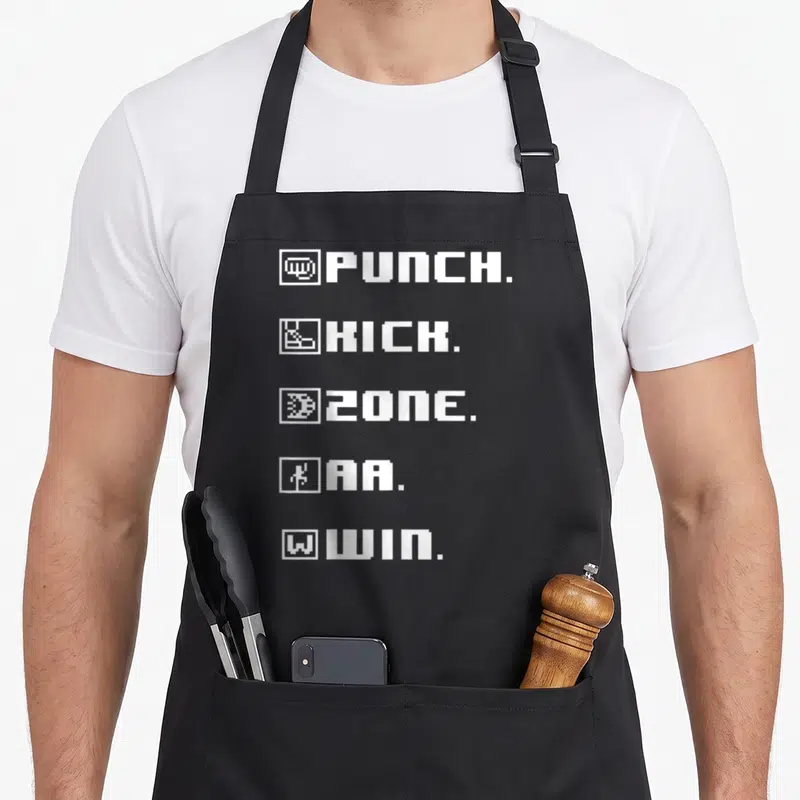 Punch game aprons - White pixel text PUNCH KICK ZONE AA WIN stacked vertically with small square icons — shown on a black cotton canvas apron against a white background