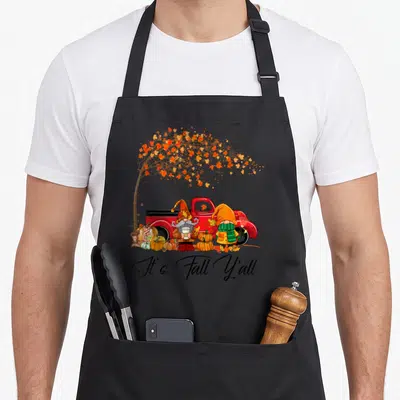 Truck fall aprons - Two gnomes near red truck with pumpkins and falling orange yellow leaves under It's Fall Y'all text — shown on a black cotton canvas apron against a white background