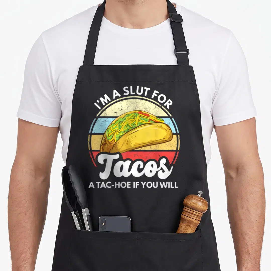Tac taco apron - Retro striped circle with taco and text I'M A SLUT FOR Tacos A TAC-HOE IF YOU WILL — shown on a black cotton canvas apron against a white background