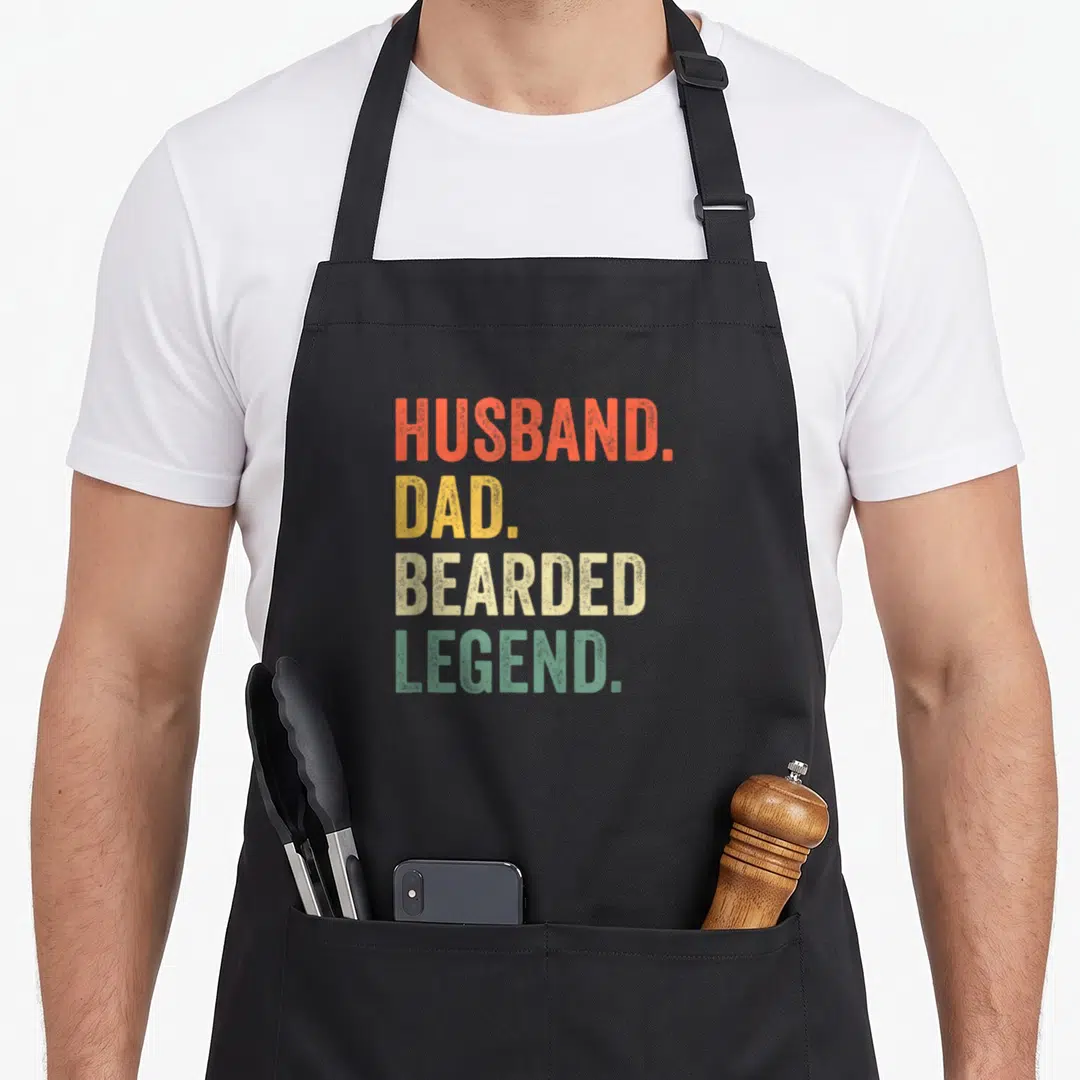 Beard father day apron - Stacked text HUSBAND DAD BEARDED LEGEND in orange red yellow beige green block letters — shown on a black cotton canvas apron against a white background