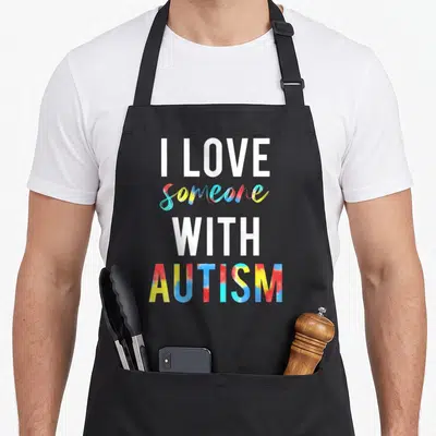 Someone teacher art apron - Stacked text I Love Someone With Autism in white and rainbow tie-dye colors — shown on a black cotton canvas apron against a white background