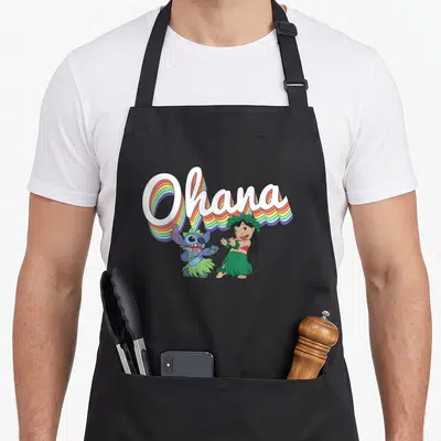 Ohana rainbow apron - White Ohana text with rainbow 3D shadows above blue character and girl in green grass skirt — shown on a black cotton canvas apron against a white background