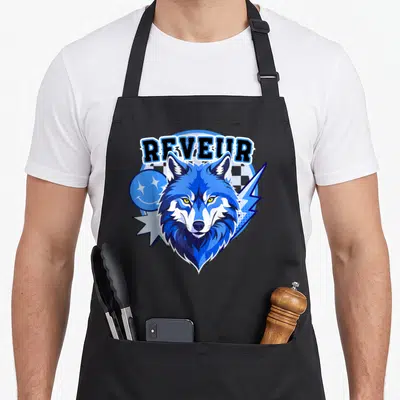 Funny dreamer apron - Blue wolf head with REVEUR text, lightning bolt, and smiley face in cartoon style — shown on a black cotton canvas apron against a white background