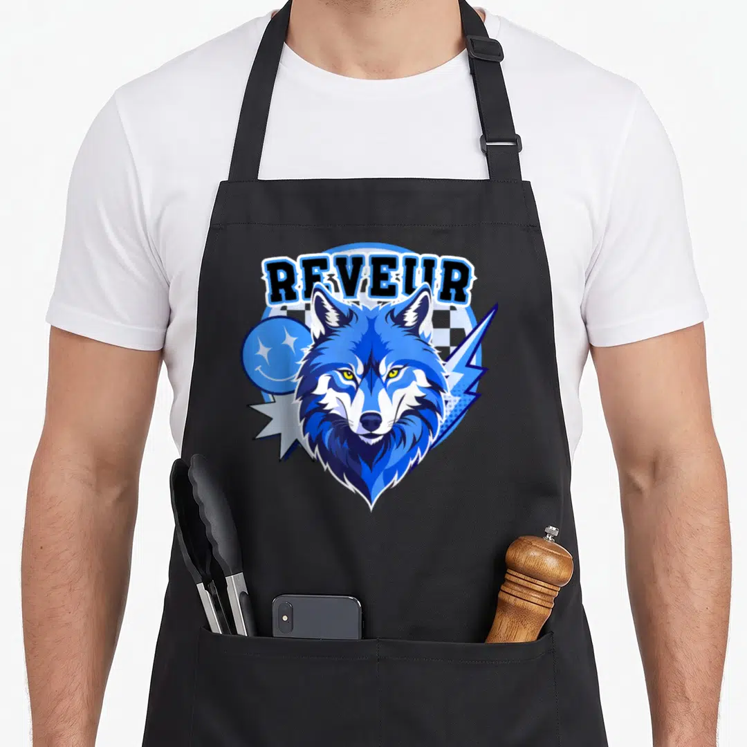 Funny dreamer apron - Blue wolf head with REVEUR text, lightning bolt, and smiley face in cartoon style — shown on a black cotton canvas apron against a white background