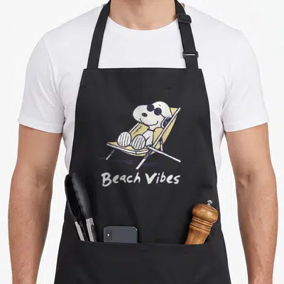 Beach family aprons - Snoopy in yellow chair with Beach Vibes text in cartoon style — shown on a black cotton canvas apron against a white background