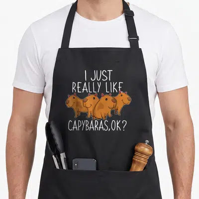 Cute capybara apron - Four brown cartoon capybaras under white text reading I Just Really Like Capybaras OK — shown on a black cotton canvas apron against a white background