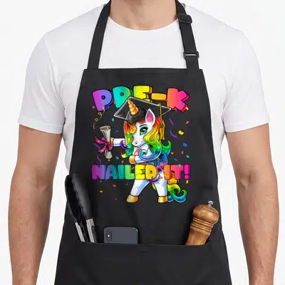 Flossing pre unicorn apron - Cartoon unicorn with rainbow mane wearing graduation cap holding diploma with PRE-K Nailed — shown on a black cotton canvas apron against a white background