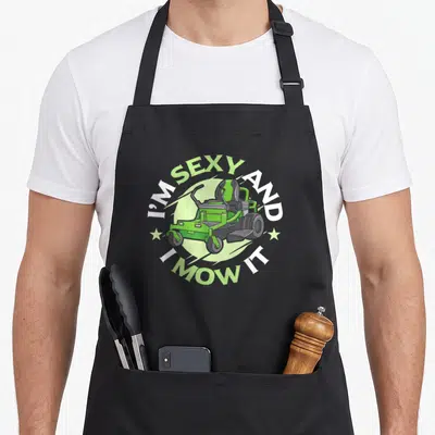 Mow service apron - Green riding mower in circle with I'M SEXY AND, I MOW IT text and two stars — shown on a black cotton canvas apron against a white background