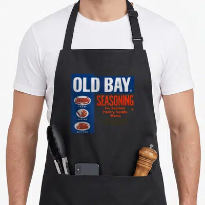 Bay spice apron - Old Bay Seasoning logo with crab, shrimp, and chicken icons in blue and red — shown on a black cotton canvas apron against a white background