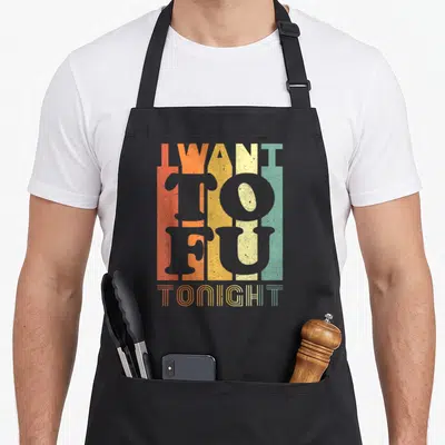 Tofu vegan apron - I WANT TO FU TONIGHT text over vertical orange yellow beige green grey stripes — shown on a black cotton canvas apron against a white background