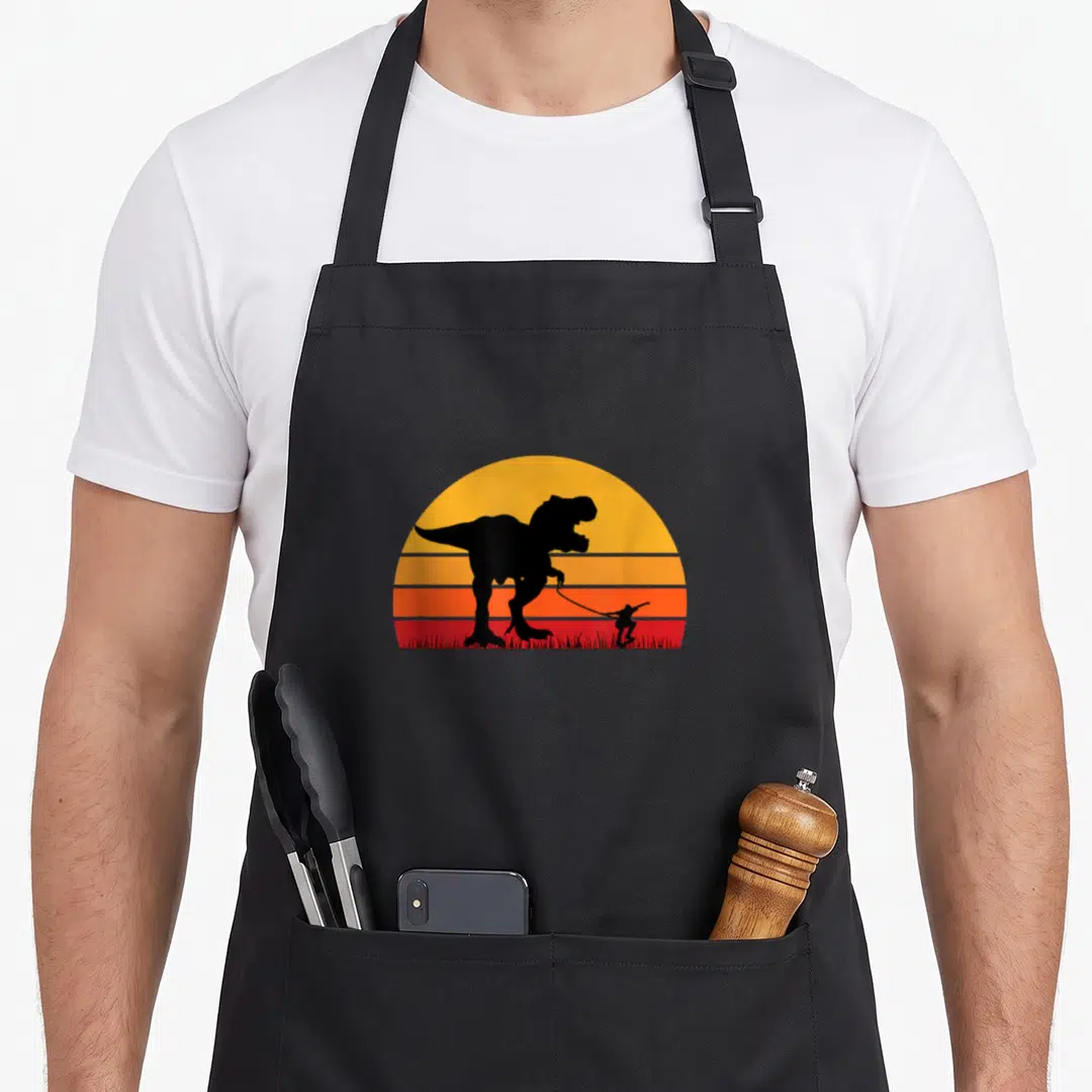Rex walking apron - Black T-Rex silhouette holding a leash attached to a skateboarder against a yellow orange and red — shown on a black cotton canvas apron against a white background