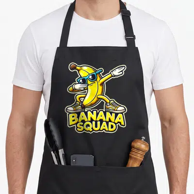 Squad banana apron - Cartoon banana in sunglasses and sneakers dabbing above stacked yellow BANANA SQUAD text — shown on a black cotton canvas apron against a white background