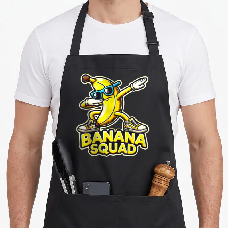 Squad banana apron - Cartoon banana in sunglasses and sneakers dabbing above stacked yellow BANANA SQUAD text — shown on a black cotton canvas apron against a white background