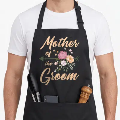 Special wedding day aprons - Beige Mother of the Groom text with pink watercolor flowers and green arrow — shown on a black cotton canvas apron against a white background