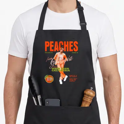 Justin peaches apron - Man in orange suit magazine cover design with barcode peach and bold typography — shown on a black cotton canvas apron against a white background