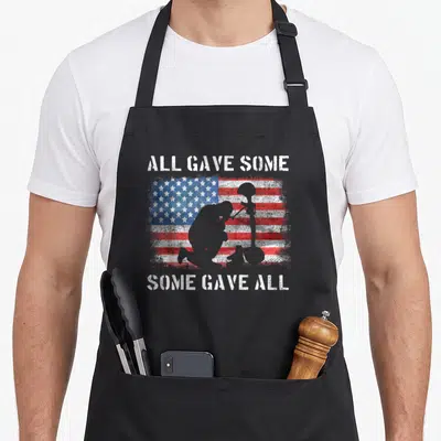 Gave veterans day apron - Kneeling soldier silhouette and helmet next to rifle over distressed red white blue flag — shown on a black cotton canvas apron against a white background