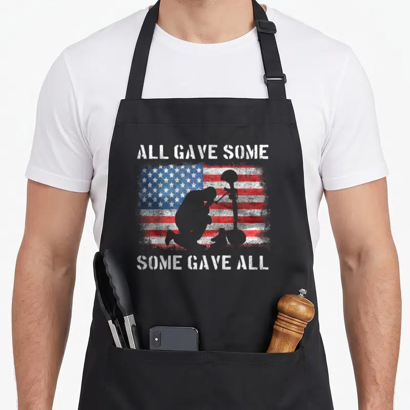 Gave veterans day apron - Kneeling soldier silhouette and helmet next to rifle over distressed red white blue flag — shown on a black cotton canvas apron against a white background