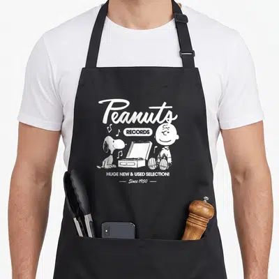 Charlie record apron - White Peanuts Records graphic with Charlie Brown, Snoopy, and turntable on black bib — shown on a black cotton canvas apron against a white background