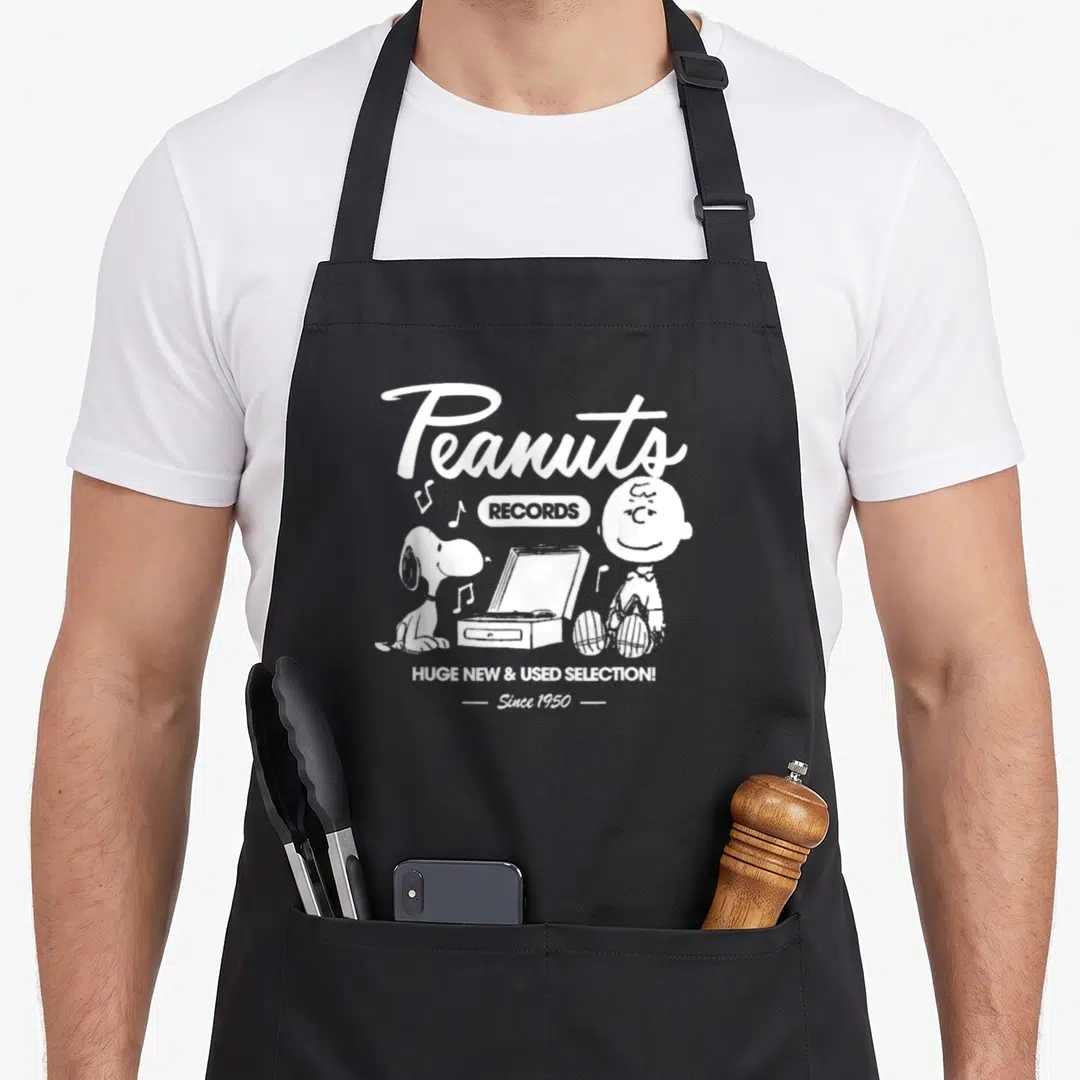 Charlie record apron - White Peanuts Records graphic with Charlie Brown, Snoopy, and turntable on black bib — shown on a black cotton canvas apron against a white background