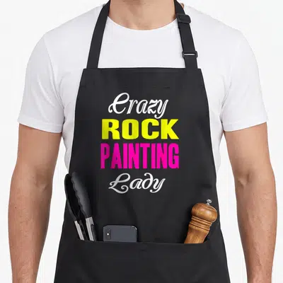 Painting novelty apron - Stacked text reading Crazy Rock Painting Lady in white, yellow, and pink fonts — shown on a black cotton canvas apron against a white background
