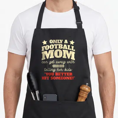 Cool football apron - Stacked beige and red text reading football mom and hit someone with a dark football graphic — shown on a black cotton canvas apron against a white background