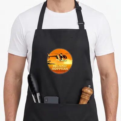 Hopper kitchen island apron - Circular logo with flying helicopter, orange sunset, palm trees, and stacked Island — shown on a black cotton canvas apron against a white background
