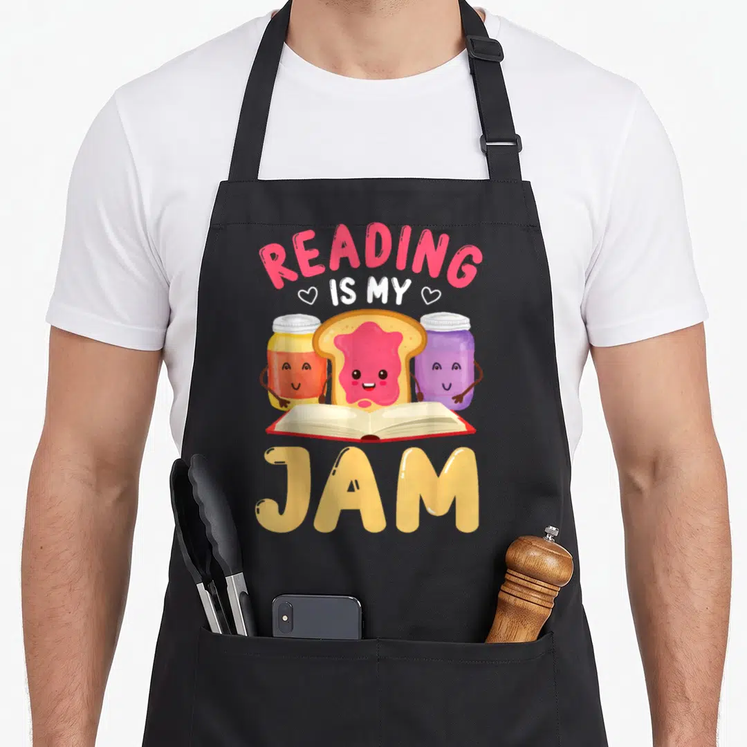 Reading meet the author apron - Cartoon toast and jam jars behind open book with arched READING IS MY JAM text in pink — shown on a black cotton canvas apron against a white background