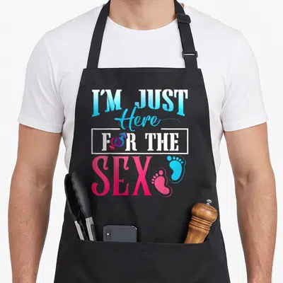 Cool gender reveal apron - Stacked text I'M JUST Here FOR THE SEX with baby footprints and gender symbol in blue pink — shown on a black cotton canvas apron against a white background