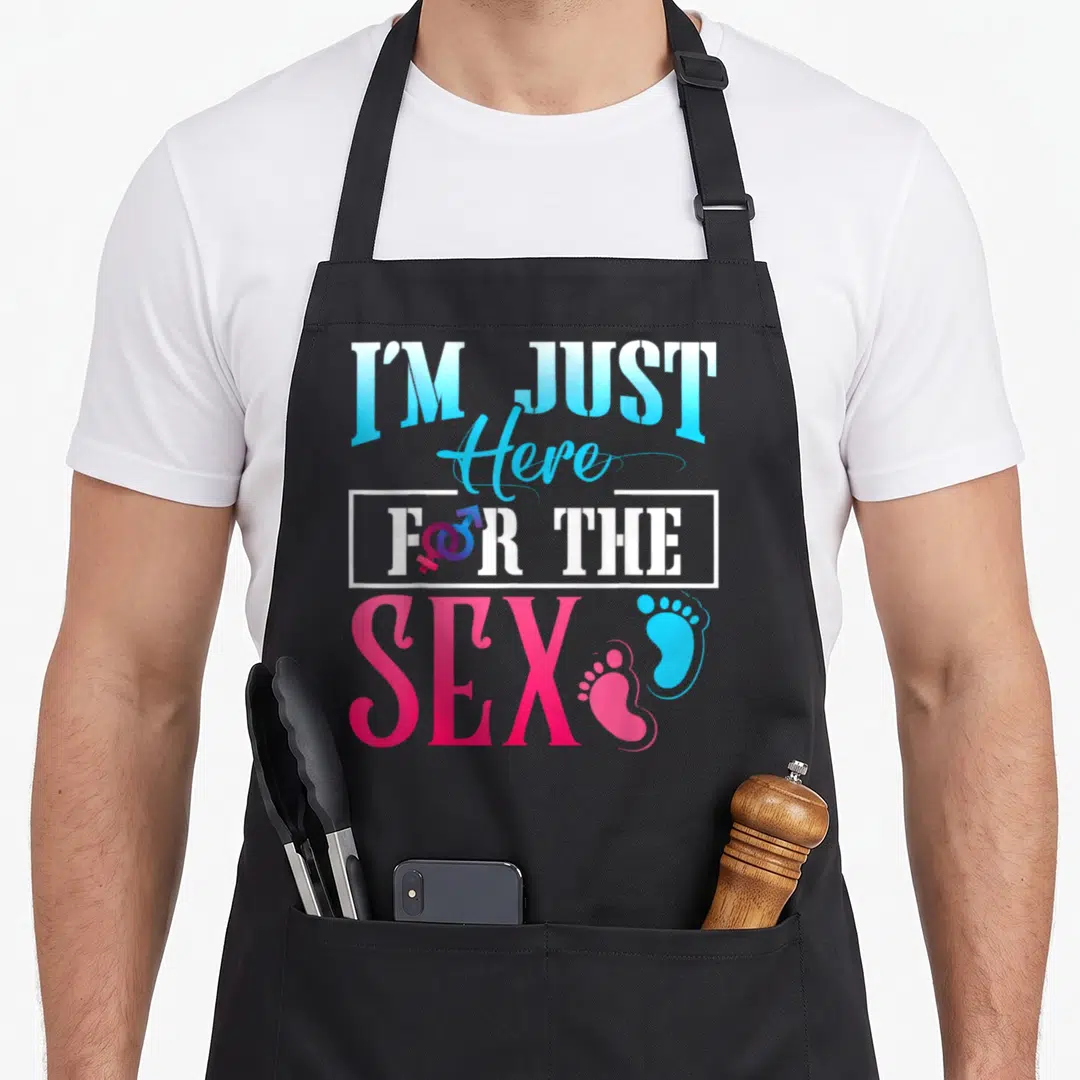 Cool gender reveal apron - Stacked text I'M JUST Here FOR THE SEX with baby footprints and gender symbol in blue pink — shown on a black cotton canvas apron against a white background
