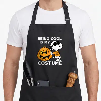 Hobby halloween apron - Snoopy in Joe Cool shirt next to orange pumpkin with white text — shown on a black cotton canvas apron against a white background