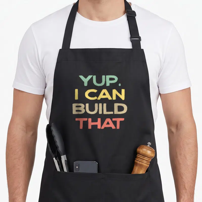 Yup build woodworking apron - Stacked text YUP I CAN BUILD THAT in green yellow beige red — shown on a black cotton canvas apron against a white background