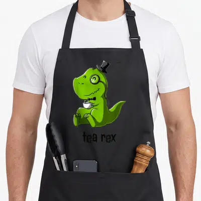 Tea coffee apron - Green cartoon T-Rex in top hat and monocle holding a white teacup with text tea rex — shown on a black cotton canvas apron against a white background
