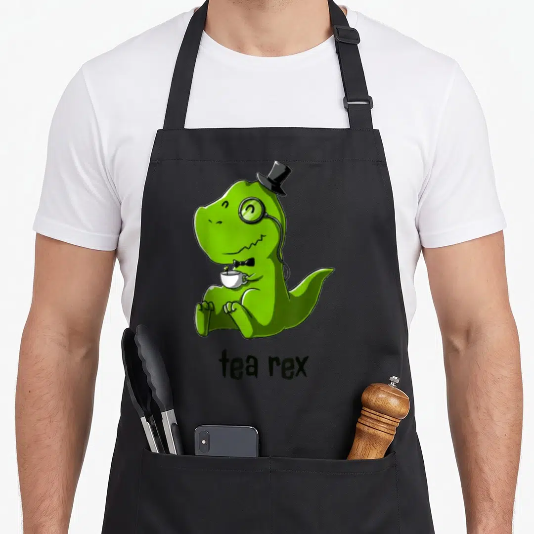 Tea coffee apron - Green cartoon T-Rex in top hat and monocle holding a white teacup with text tea rex — shown on a black cotton canvas apron against a white background