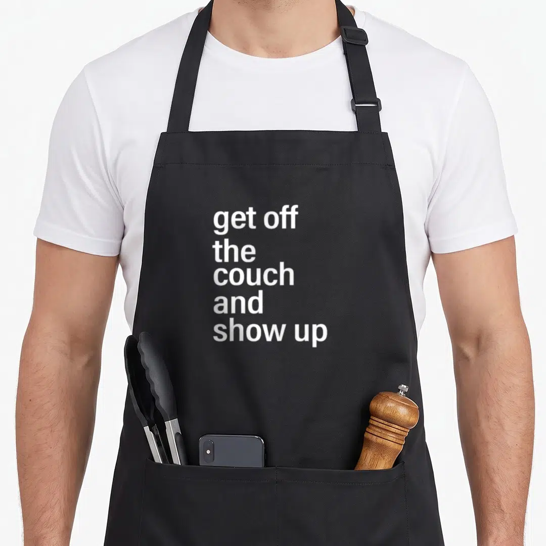 Couch jd apron - white stacked text reading get off the couch and show up — shown on a black cotton canvas apron against a white background