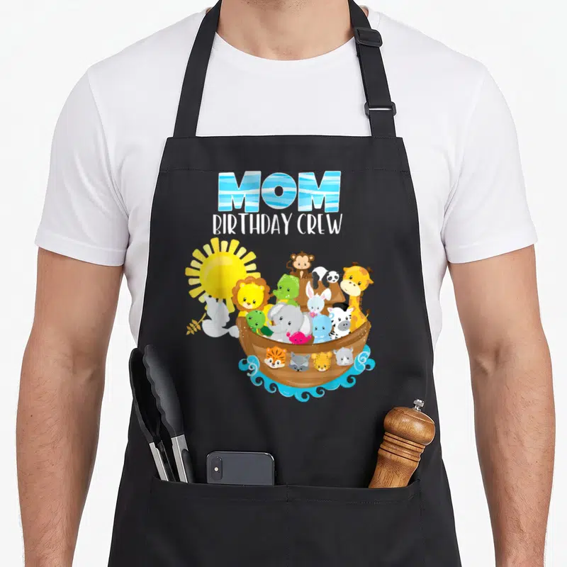Noah birthday apron - MOM BIRTHDAY CREW text above cartoon ark with animals, sun, and flying dove on blue waves — shown on a black cotton canvas apron against a white background