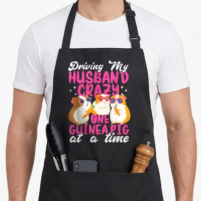 Driving husband wife aprons - Three cartoon guinea pigs with sunglasses holding carrots under stacked pink and white — shown on a black cotton canvas apron against a white background