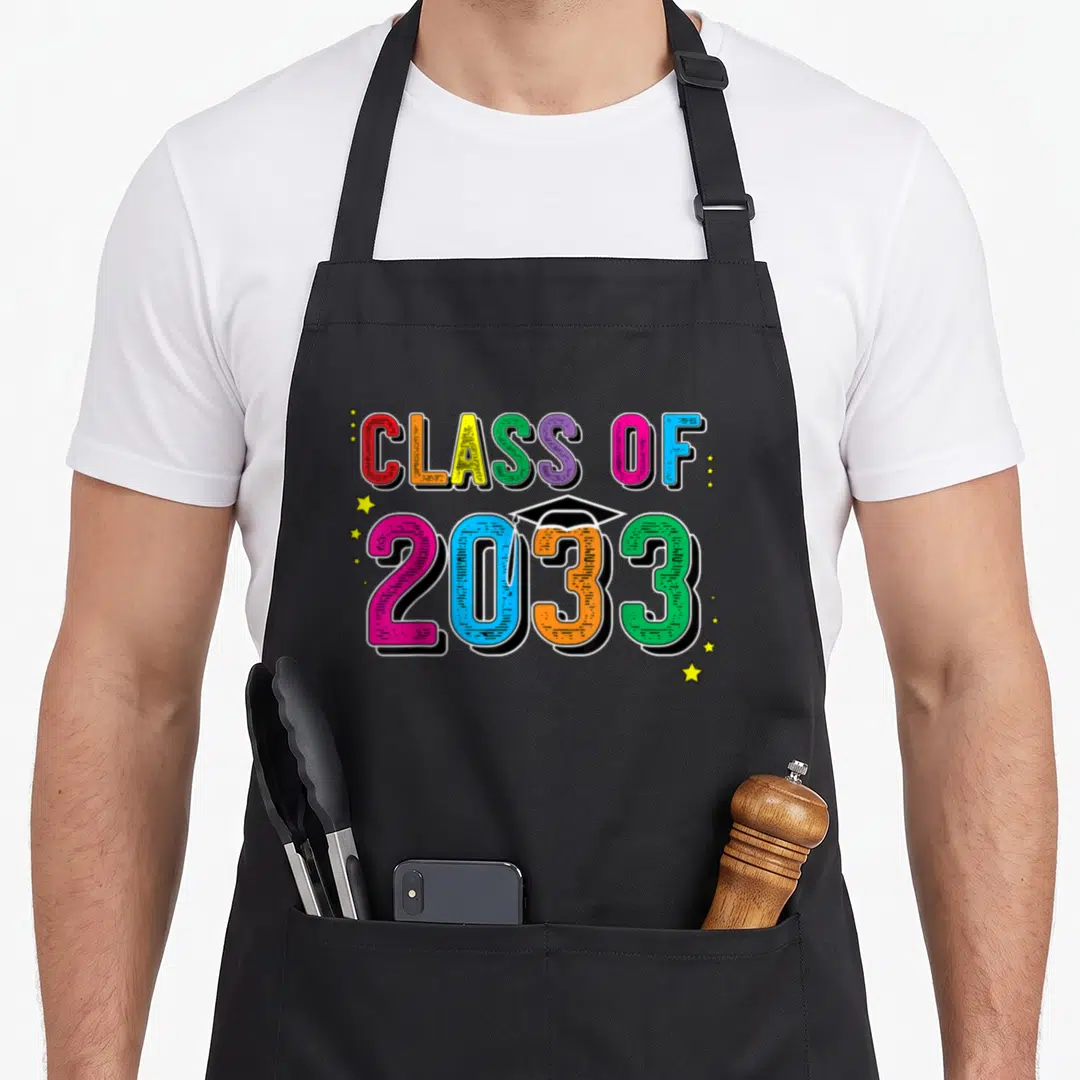 School class apron - Class of 2033 rainbow text with graduation cap and yellow stars — shown on a black cotton canvas apron against a white background